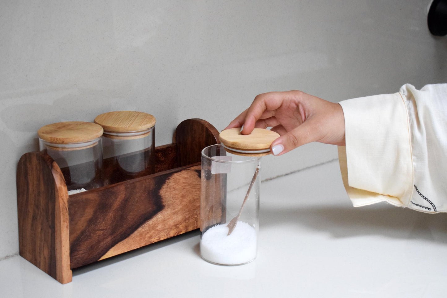 Hand reaching for a jar lid next to a wooden container set on a light surface.
