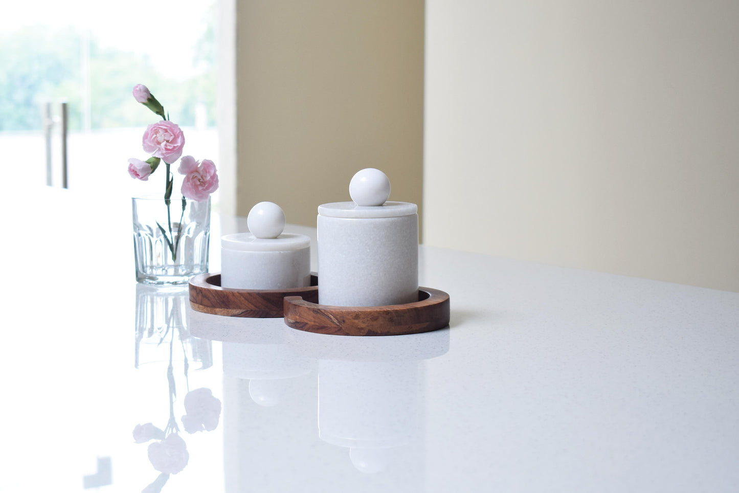 Two white cylindrical marble food jars with wooden tray, with pink flowers in the background.