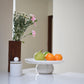 Fruits on a white fruit stand with a vase of flowers in the background